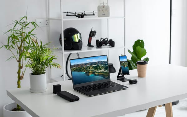 A tabletop with gadgets, laptops, plants, and a bookcase with a helmet, rc car, drone and equipment